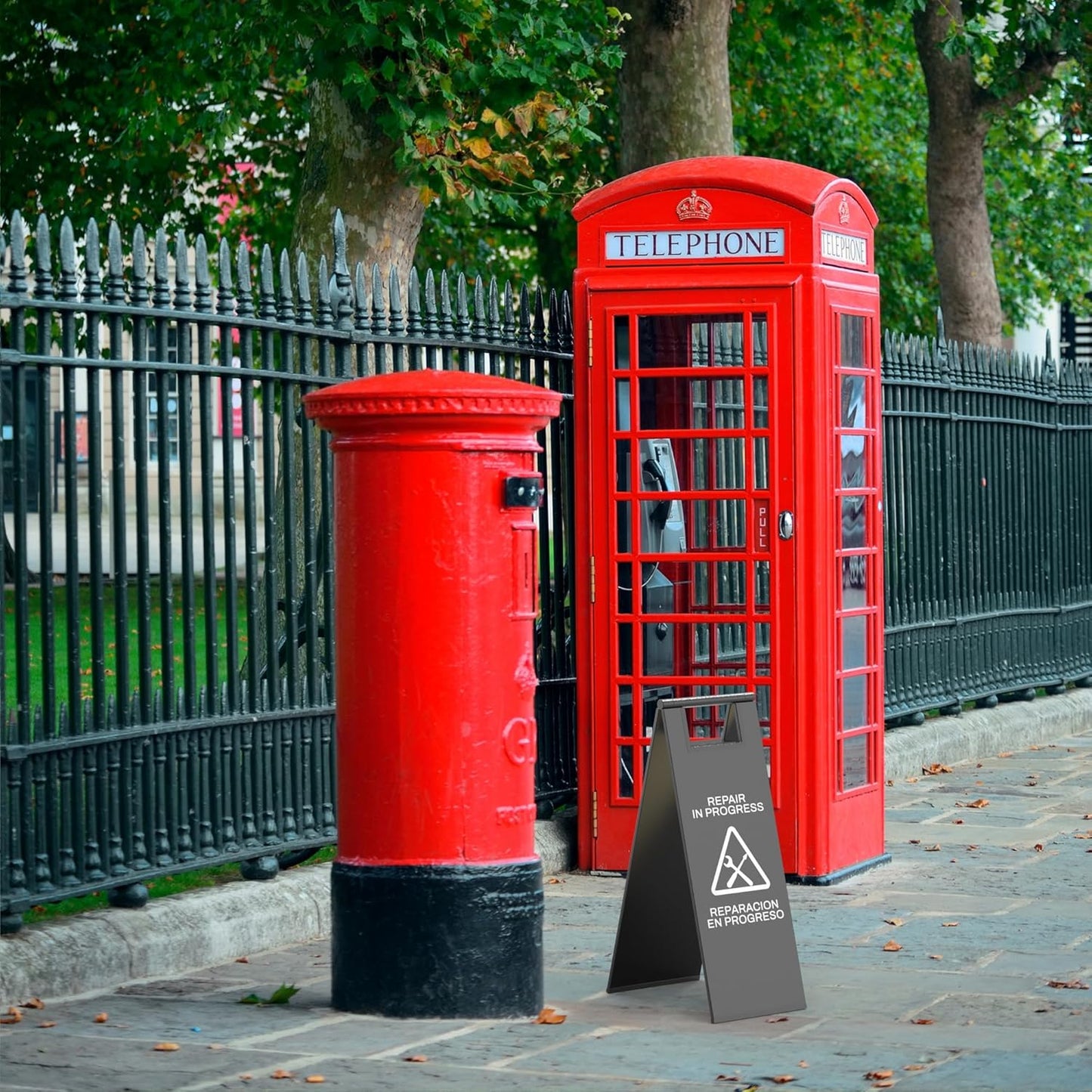 Black public facility repair sign with triangular warning placed next to red British telephone box during maintenance
