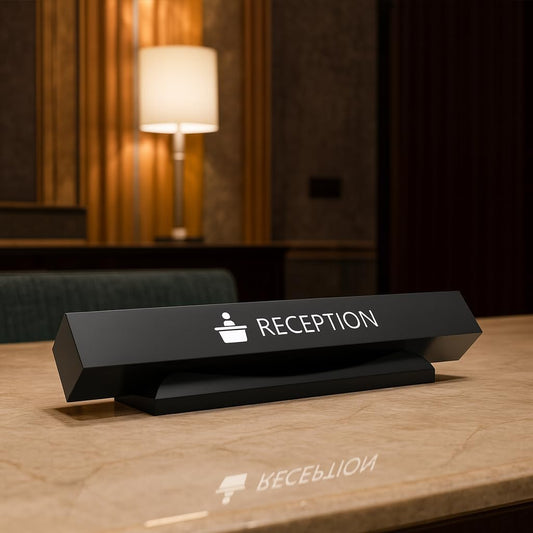 Elegant black triangular reception desk sign with white text and icon, positioned on wooden hotel reception counter with warm ambient lighting
