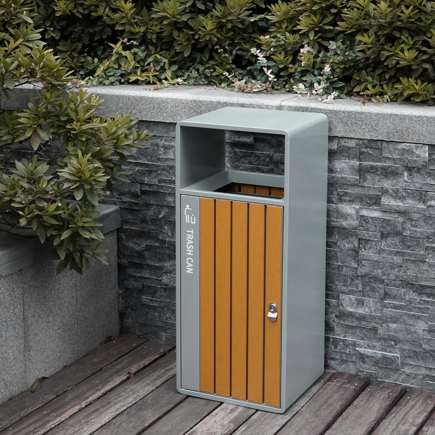 Heavy-duty steel outdoor trash can with natural wood panel door, positioned on wooden deck beside stone wall and greenery
